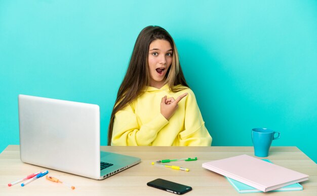 Little girl in a table with a laptop over isolated blue background surprised and pointing side