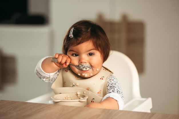 A little girl todler eats porridge herself with a spoon. first feeding