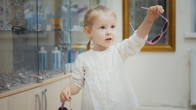 Little girl tries fashion medical glasses near mirror - shopping in ophthalmology clinic, close up