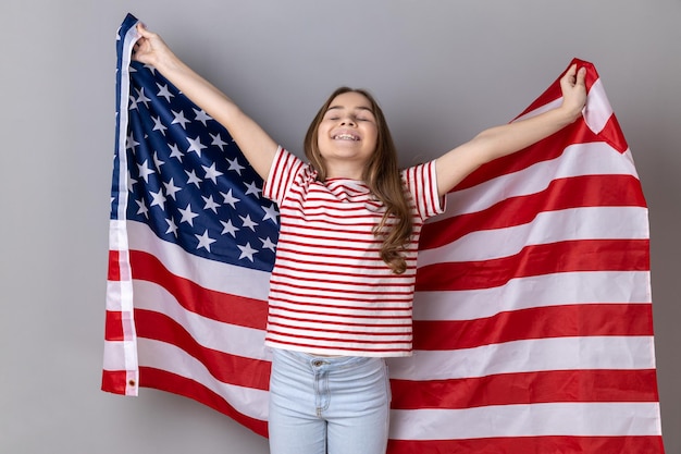 T셔츠를 입은 어린 소녀가 어깨에 미국 국기를 들고 눈을 감고 행복하게 웃고 있습니다