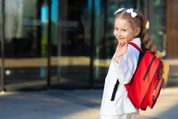 La bambina in uniforme va in prima elementare, agitando la mano. bambino che ride felice torna a scuola. ragazzo con lo zainetto rosso nel campus.