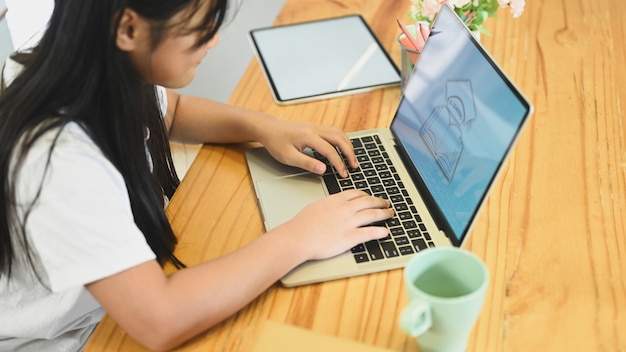 A little girl uses a computer laptop at a wooden working desk. Studying at home, E-learning concept.