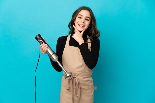 Little girl using hand blender isolated on blue wall happy and smiling