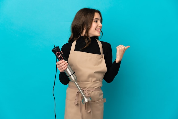 Little girl using hand blender isolated on blue wall pointing to the side to present a product