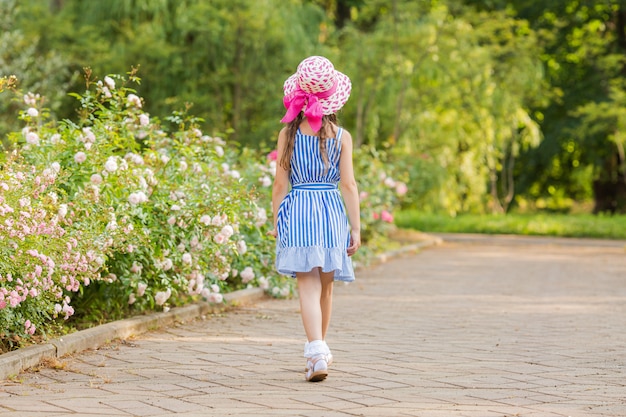 Little girl walks in the garden with blooming roses. stands back in a hat with a pink ribbon, space for text