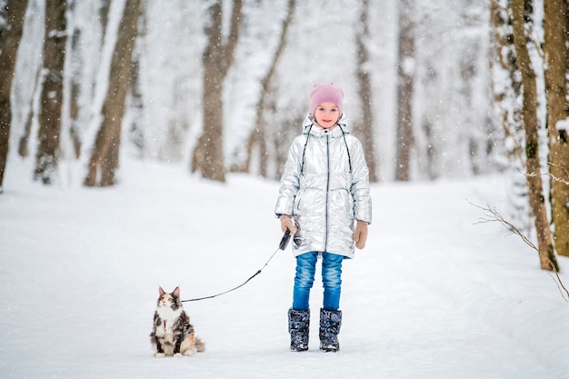 小さな女の子は雪に覆われたウィンターパークでひもにつないで猫と一緒に歩く