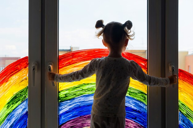 Little girl on wall of painting rainbow on window. Kids leisure at home. Positive visual support during quarantine Pandemic Coronavirus Covid-19 at home.