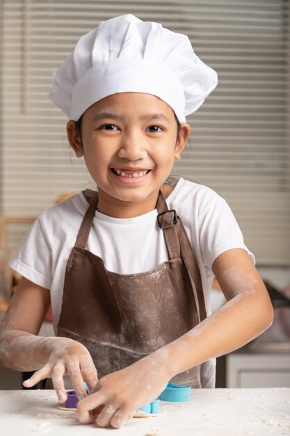 Маленькая девочка надевает белую шляпу шеф-повара и коричневый фартук для приготовления печенья
