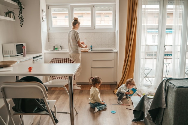 Little girl watching her sister drawing with a pencil on the\
paper while their female parent cooking breakfast