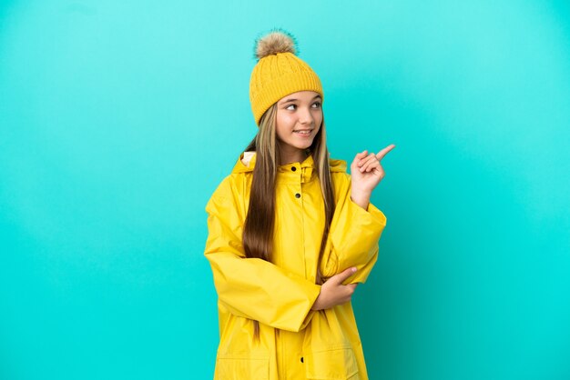 Фото Маленькая девочка в непромокаемом пальто на изолированном синем фоне указывает на отличную идею
