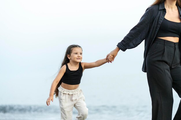 Маленькая девочка в черном топ-костюме и бежевых брюках держит маму за руку, гуляя по пляжу и веселясь.