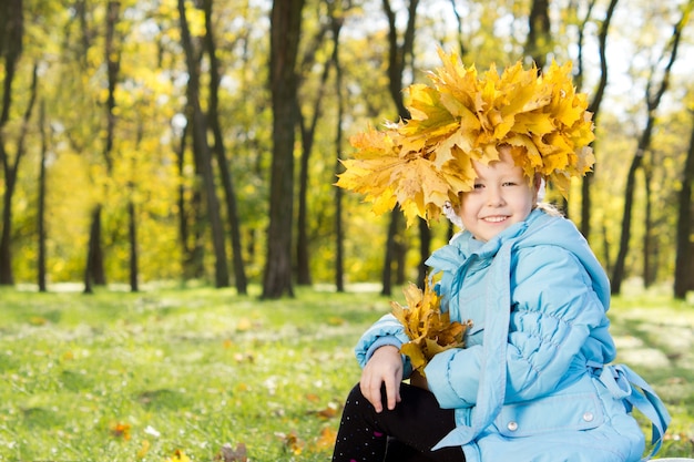 Маленькая девочка в короне из разноцветных желтых осенних листьев на голове, счастливо улыбаясь, в осеннем лесу