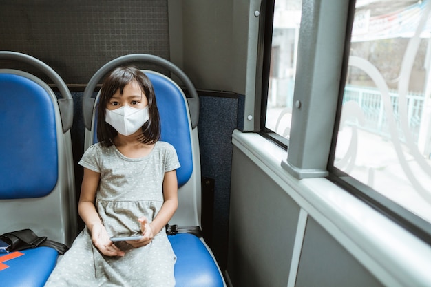 A little girl wearing a mask sits on a bench holding a cellphone on the bus while traveling