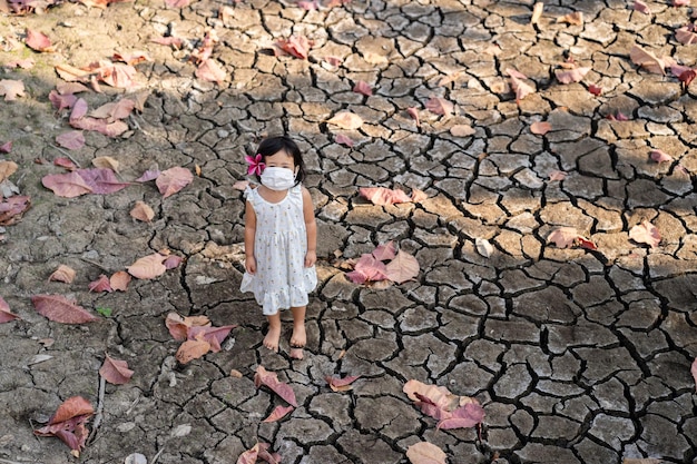 Foto piccola ragazza che indossa una maschera medicale in piedi presso uno stagno secco durante la stagione secca