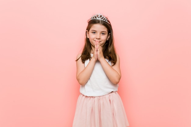 Little girl wearing a princess look holding hands in pray near mouth, feels confident.