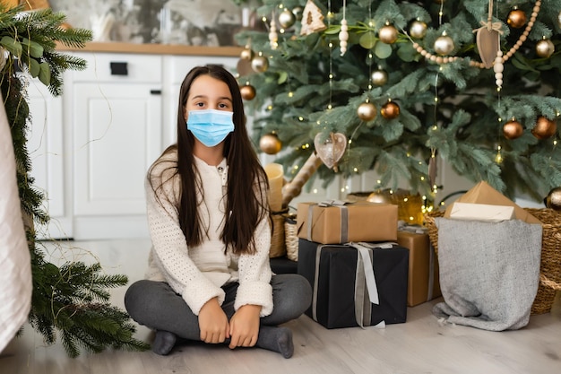 Little girl wearing protective mask on Christmas tree background. Sad child face with surgical mask. Christmas festive and social distancing during covid-19 pandemic concept.