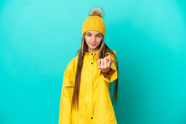 来るジェスチャーをしている孤立した青い背景の上に防雨コートを着ている少女