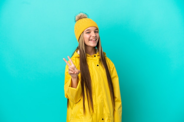 Little girl wearing a rainproof coat over isolated blue background smiling and showing victory sign