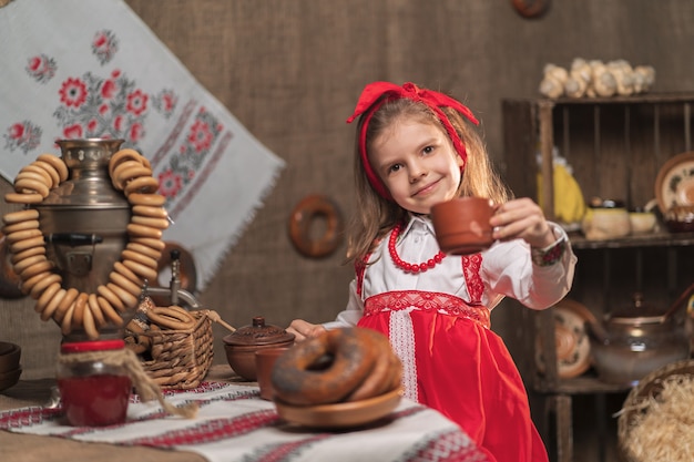 Фото Маленькая девочка в красной повязке и декоративной рубашке пьет чай из самовара, празднуя масленицу