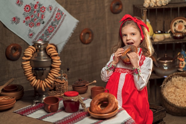 Little girl wearing red headband and ornamental shirt sitting at table full of food and big samovar celebrating Maslenitsa