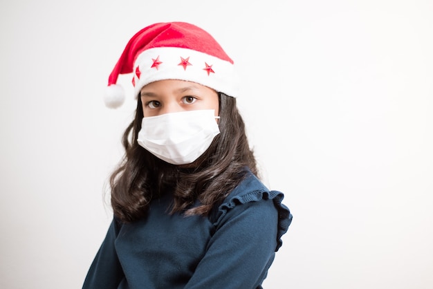 Little girl wearing santa hat and face mask looking to the camera sad on white background.