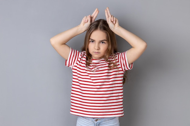 Little girl wearing striped Tshirt showing bull horns gesture over head frowning as before attack