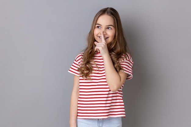 Little girl wearing striped Tshirt standing showing hush sign and looking away with toothy smile