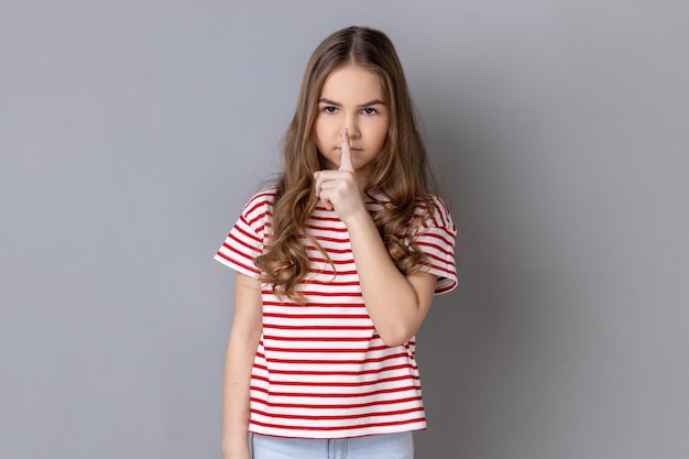 Tシャツを着た小さな女の子が口に指を当てて沈黙のジェスチャーを示し、静かにしているように頼む