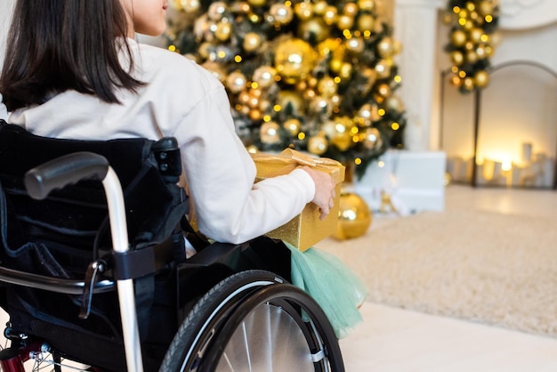 크리스마스 트리 근처 휠체어에 어린 소녀입니다. 노란색, 황금색 새해 선물을 들고 있는 장애인