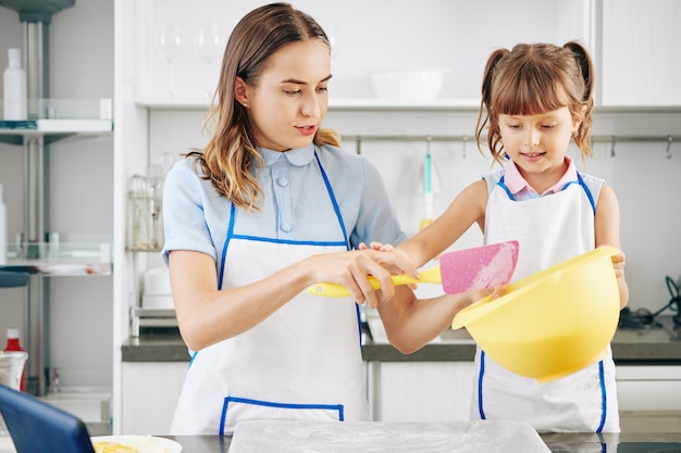 Little girl in white apron helping mother with making cookie dough and baking in kitchen