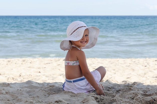 Little girl in a white beach hat and bikini sits on the sand by the sea