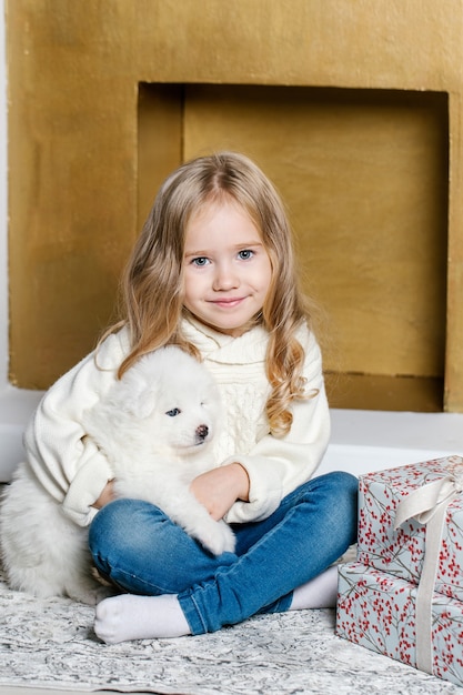 Little girl in white clothes are sitting on the floor near the tree with white Samoyed puppy and smiling.