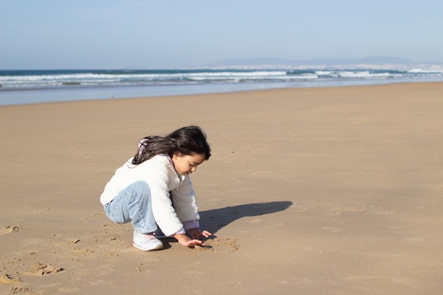 Little girl in white coat playing on beach alone. Japanese child drawing on wet sand on summer day. Childhood, portrait, happiness concept