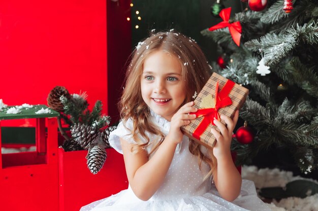 A little girl in a white dress is sitting on the floor and holding a New Year's gift against the background of a Christmas tree the concept of Christmas and new year