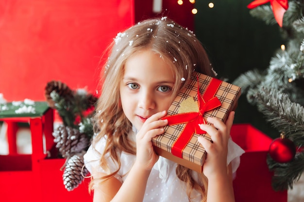 A little girl in a white dress is sitting on the floor and holding a New Year's gift against the background of a Christmas tree the concept of Christmas and new year