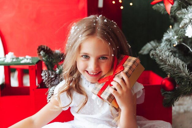 A little girl in a white dress is sitting on the floor and holding a New Year's gift against the background of a Christmas tree the concept of Christmas and new year