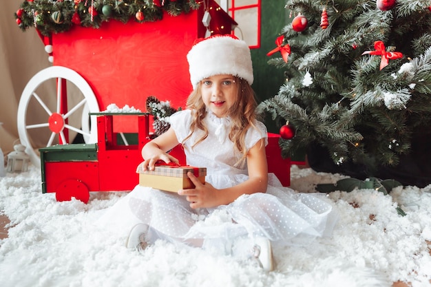 A little girl in a white dress is sitting on the floor and holding a New Year's gift against the background of a Christmas tree the concept of Christmas and new year