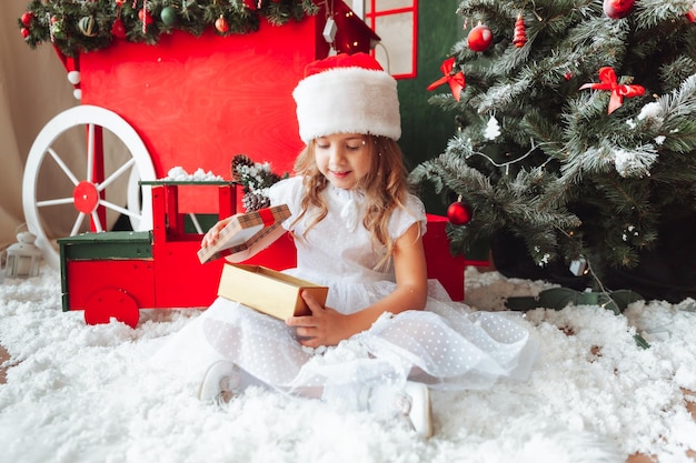 A little girl in a white dress is sitting on the floor and holding a New Year's gift against the background of a Christmas tree the concept of Christmas and new year