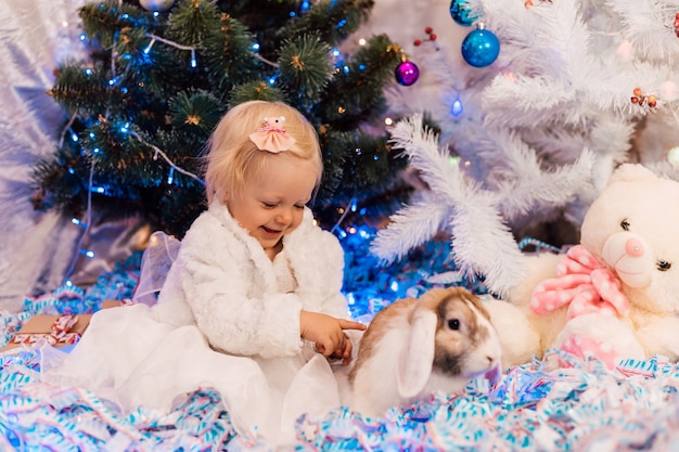 Little girl in white dress plays near the Christmas tree with a rabbit