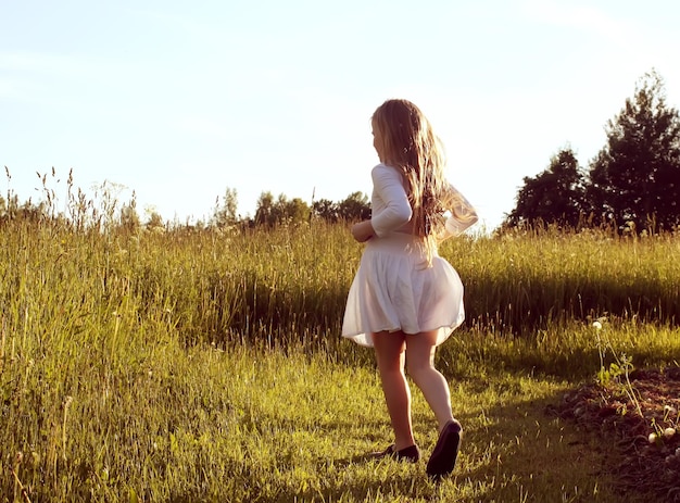 Little girl in white dress walking outdoors on the green summer field in the countryside