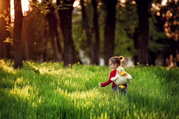 Photo little girl in white glasses walks on green meadow with duckshaped childrens backpack