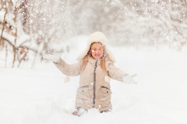 Little girl in a white hat and jacket in the winter forest throws snow. Winter fun