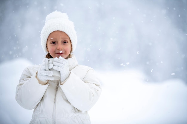 A little girl in a white jacket and a white hat is holding a cup of tea in her hands during a snowfall against the backdrop of beautiful snowdrifts and forests