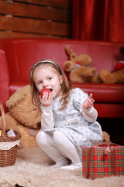 Foto bambina in abito bianco a pois mangia mela e divertirsi nello studio di natale. albero di natale, orsacchiotto e cesto con doni sul davanti.