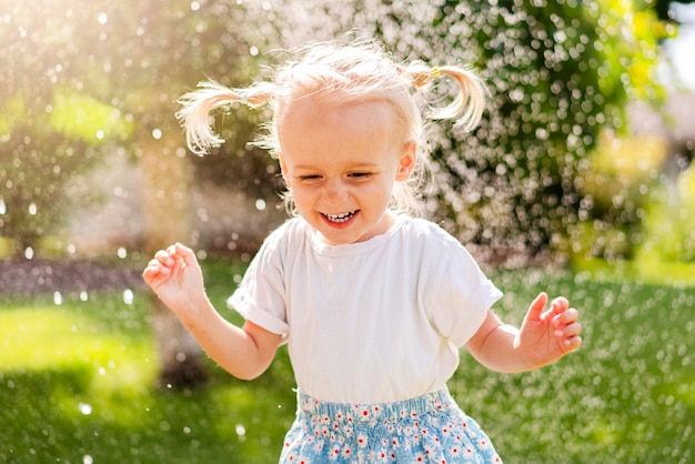 Photo a little girl in a white shirt and blue skirt runs through a sprinkler of water.