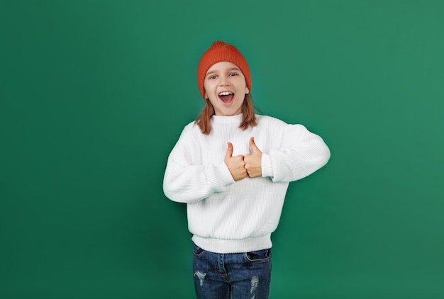 흰 스웨터와 빨간 모자를 쓴 어린 소녀가 웃고 녹색 외딴 배경에 엄지손가락을 보여줍니다.