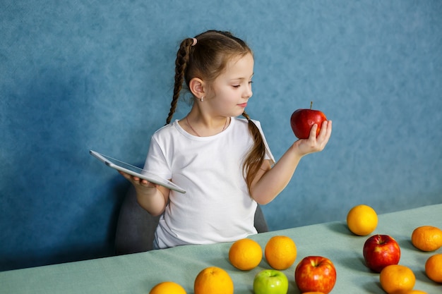 Маленькая девочка в белой футболке держит в руках планшет и изучает фрукты