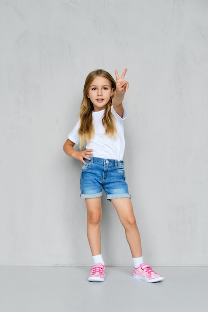 Little girl in white t-shirt, jeans shorts and pink sneakers standing shows V sign