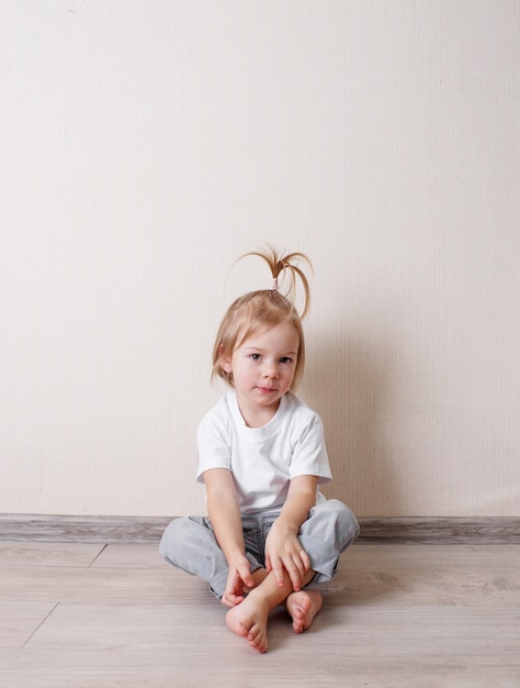 흰색 티셔츠를 입은 어린 소녀가 벽에 기대어 방 바닥에 앉아 있습니다.