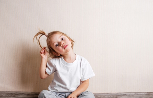 A little girl in a white t-shirt sits on the floor of the room against the wall.